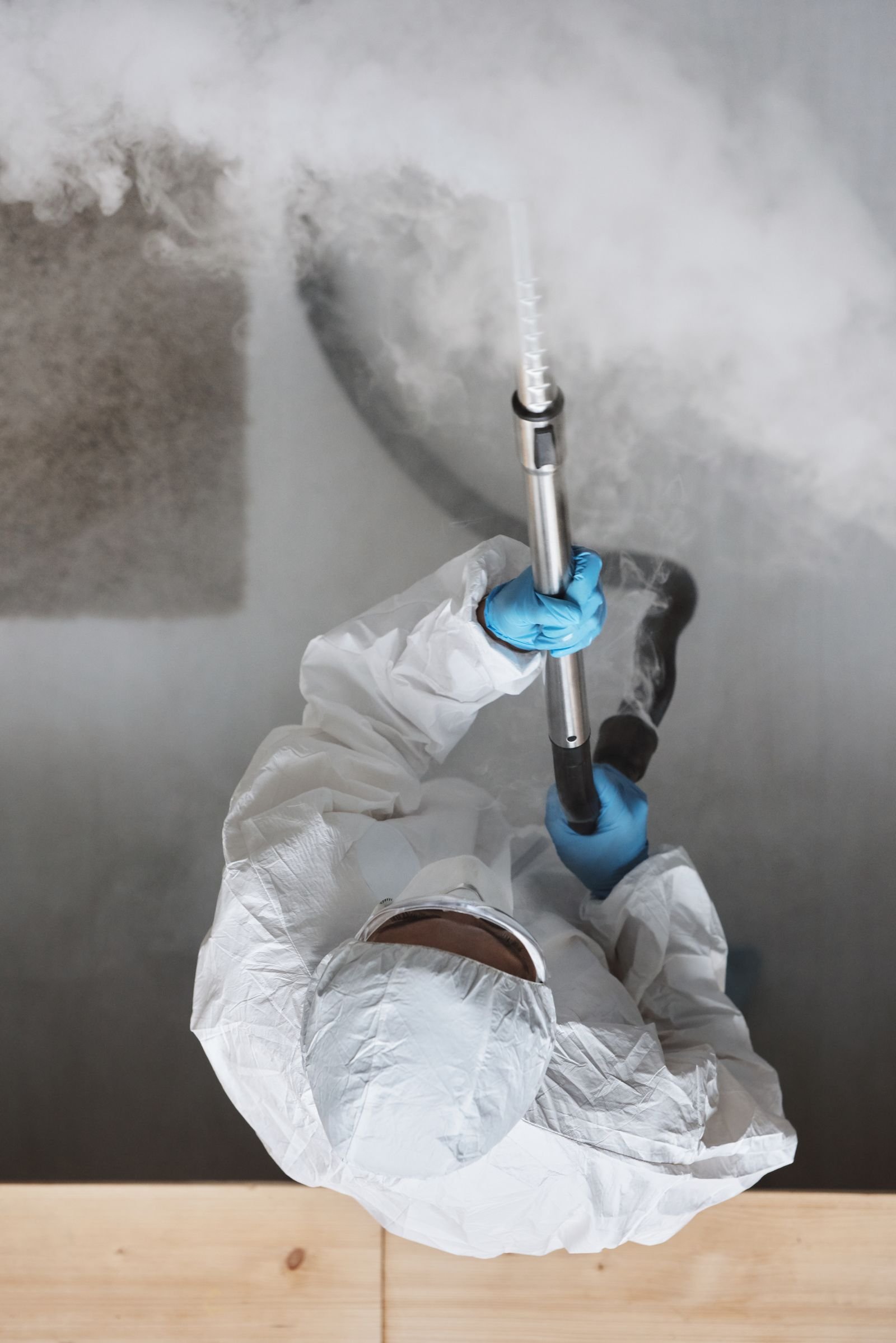 Vue plongeante sur un opérateur en combinaison blanche et gants bleus, tenant une lance de nébulisation dont sort un brouillard dense