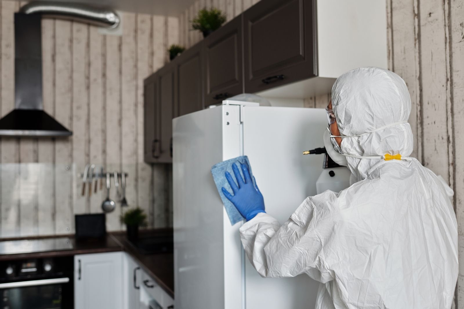 Opérateur en combinaison à capuche, masque et gants bleus, traitant la porte d’un réfrigérateur dans une cuisine inoccupée